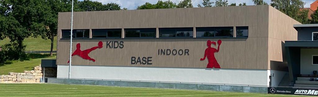 Die KIB-Arena von außen, hellbraune Fassade mit roten Spielersilhouetten hinter einem grünen Fußballplatz.