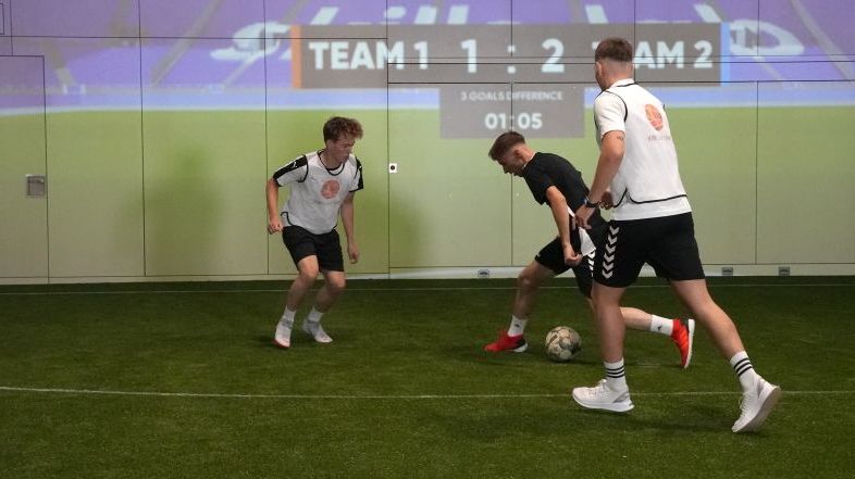 Drei junge Männer spielen in einer Indoor-Soccer-Halle gegeneinander Fußball.