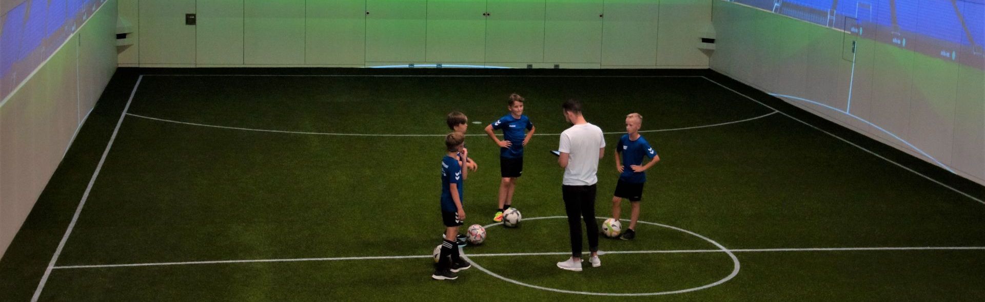 Vier Kinder und ein Trainer stehen inmitten eines Indoor-Spielfeldes und besprechen eine Übung.