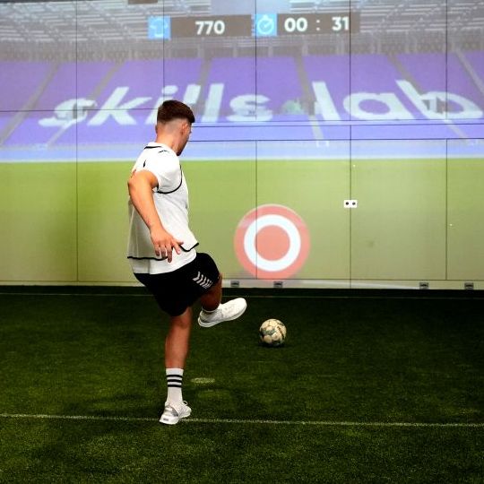 Ein junger Fußballer zielt mit dem Ball auf eine an die Wand projizierte Zielscheibe.