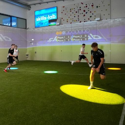 Mehrere junge Männer trainieren in  einer Indoor Soccer Halle, indem sie farbigen Lichtpunkten nachrennen.