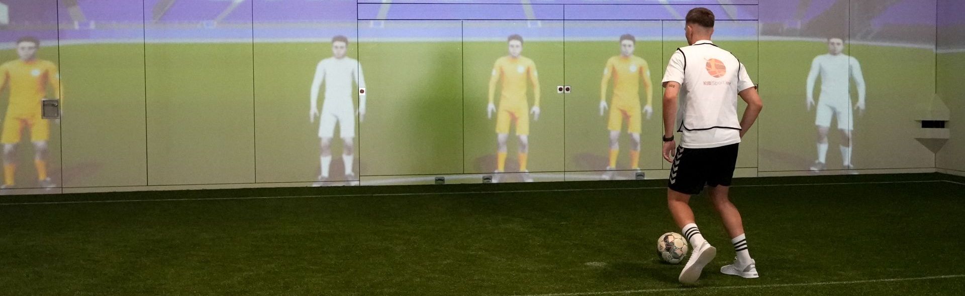 Ein junger Mann steht mit Ball vor einer Wand, an der per Projektion digitale Mit- und Gegenspieler dargestellt werden.